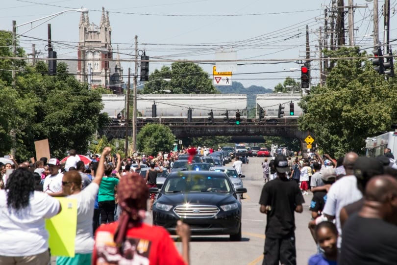 Crowds of people gather alongside roadway as hearse approaches camera