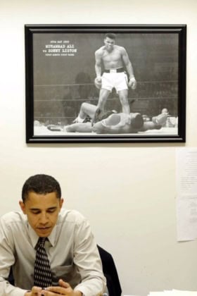 President Obama in button-up shirt and tie sitting at desk with Muhammad Ali poster behind him