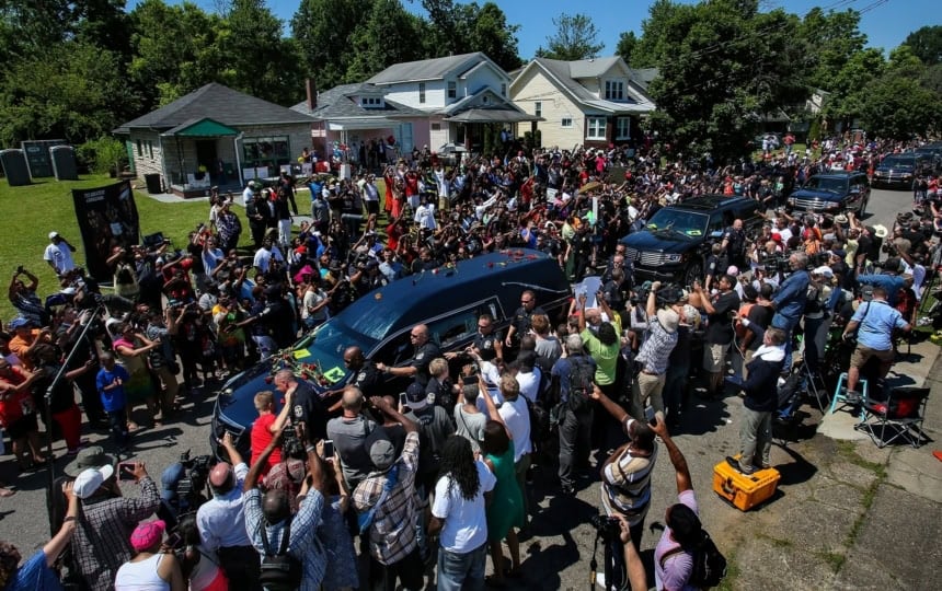Large group of people standing in roadway around black hearse