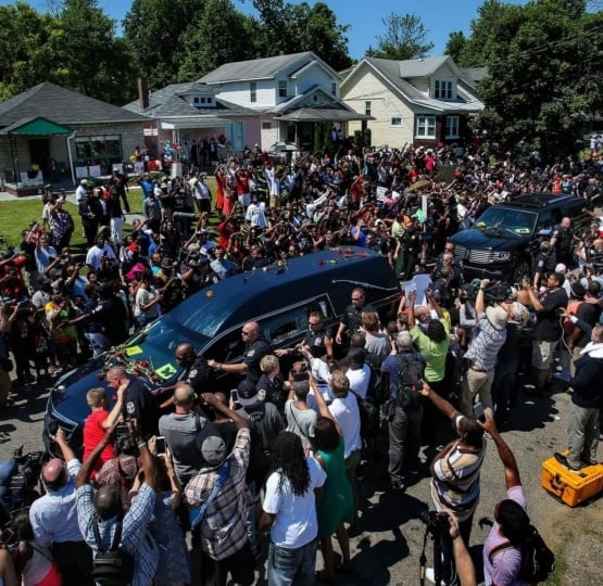 Large group of people standing in roadway around black hearse