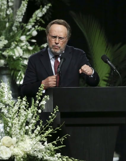 Billy Crystal speaking into a mic holding up one hand into a fist at a podium