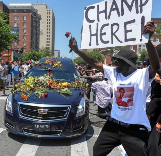 Black hearse covered in flowers with man holding up sign "Champ is Here!"