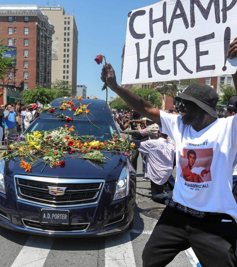 Black hearse covered in flowers with man holding up sign "Champ is Here!"