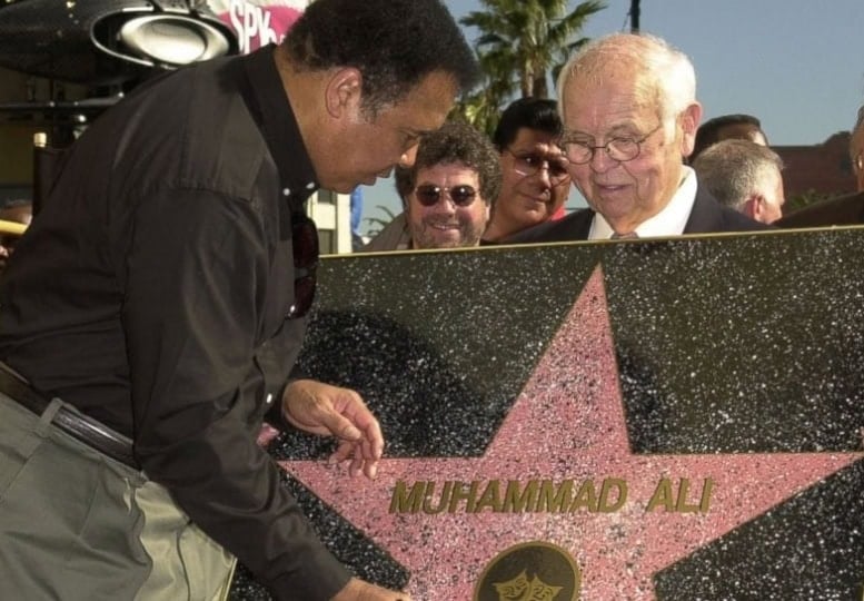 Older man holding up Hollywood Walk of Fame star while Muhammad Ali in button up shirt and gray pants signs with marker