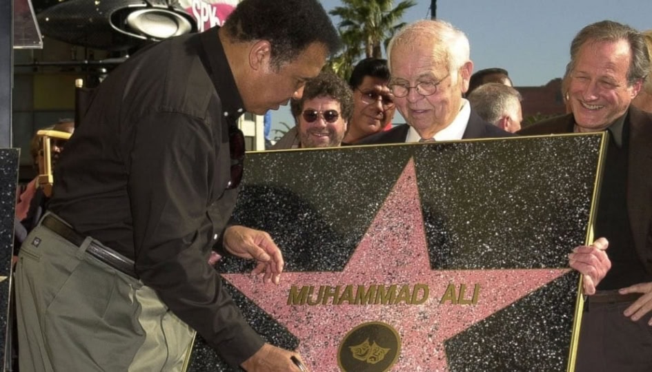 Older man holding up Hollywood Walk of Fame star while Muhammad Ali in button up shirt and gray pants signs with marker
