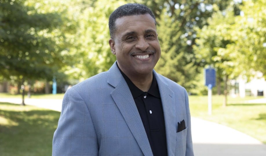 Man with short gray hair and gray moustache wearing light gray blazer and button up shirt smiling at camera in park