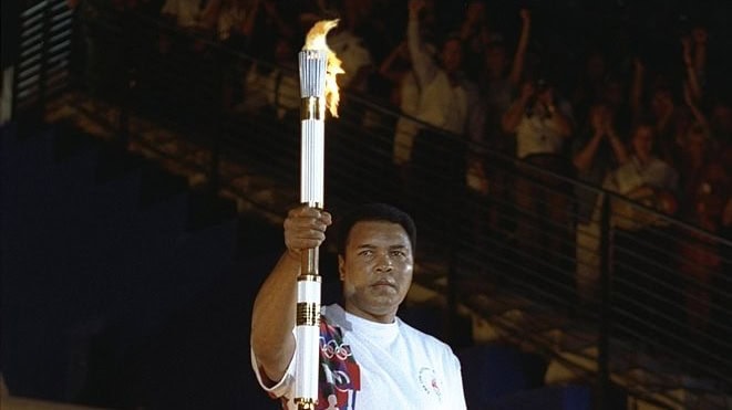 Muhammad Ali dressed in all white lifts up a lit Olympic Torch