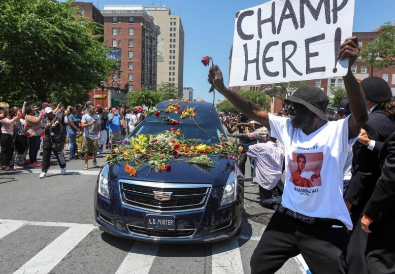 Black hearse covered in flowers with man holding up sign "Champ is Here!"