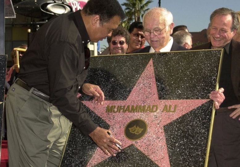 Older man holding up Hollywood Walk of Fame star while Muhammad Ali in button up shirt and gray pants signs with marker