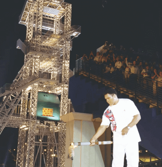 Muhammad Ali standing on stage with Olympic torch and cauldron in the background as fans cheer him pon
