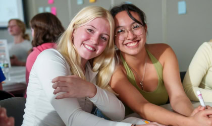 Two students smiling at a table leaning against each other