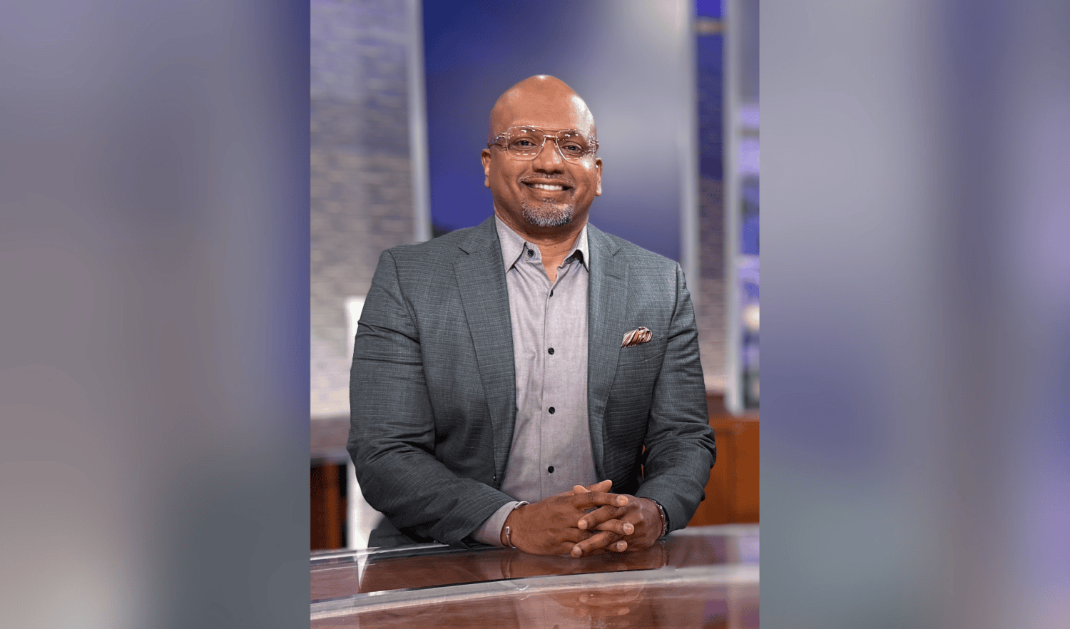 Photo of man wearing gray suit and glasses sitting at clear news desk and smiling at camera