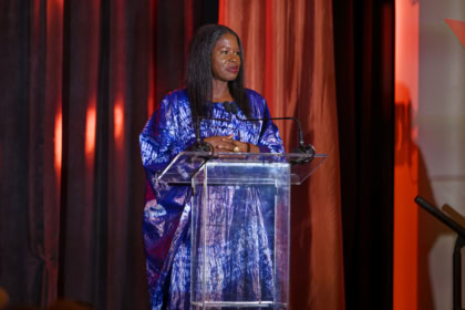 Woman with dark hair wearing shiny blue dress speaking at podium