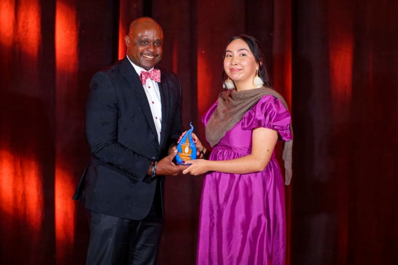 Man wearing black suit and bright bow tie giving award to woman in pink dress