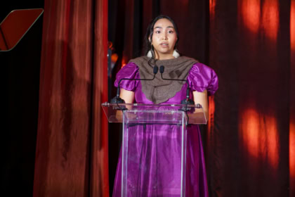 Woman with dark hair wearing earrings and purple dress speaking at podium