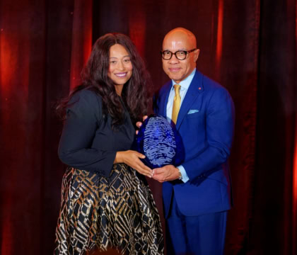 Woman with short brown hair wearing dark blue dress and man with dark glasses and light blue suit hold award and smile at camera