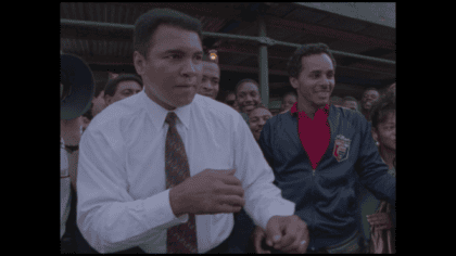 Muhammad Ali in white button-down shirt and tie in boxing pose standing among crowd in the street