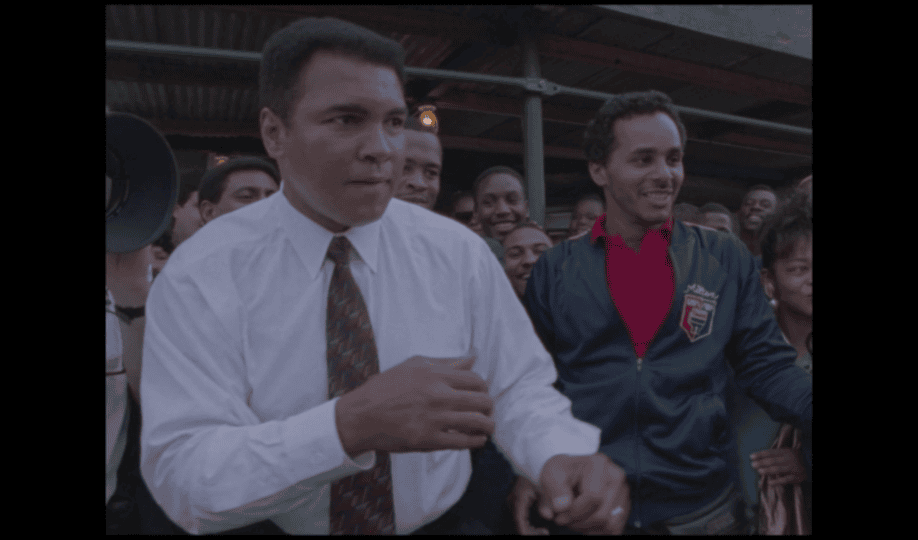 Muhammad Ali in white button-down shirt and tie in boxing pose standing among crowd in the street