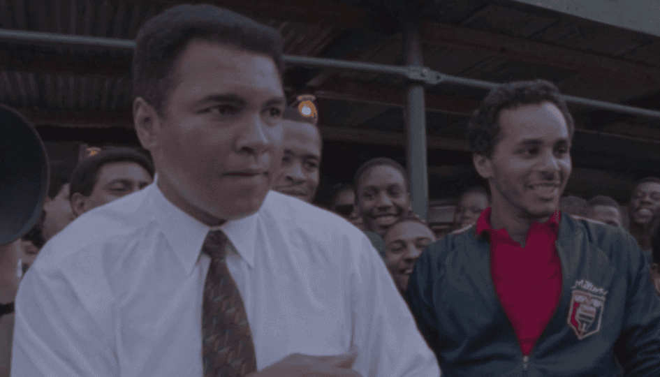 Muhammad Ali in white button-down shirt and tie in boxing pose standing among crowd in the street