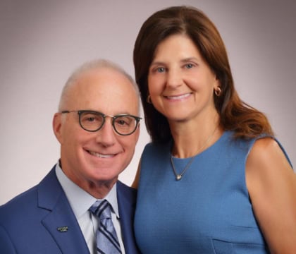Man wearing blue suit and glasses next to woman with blue dress smiling at camera